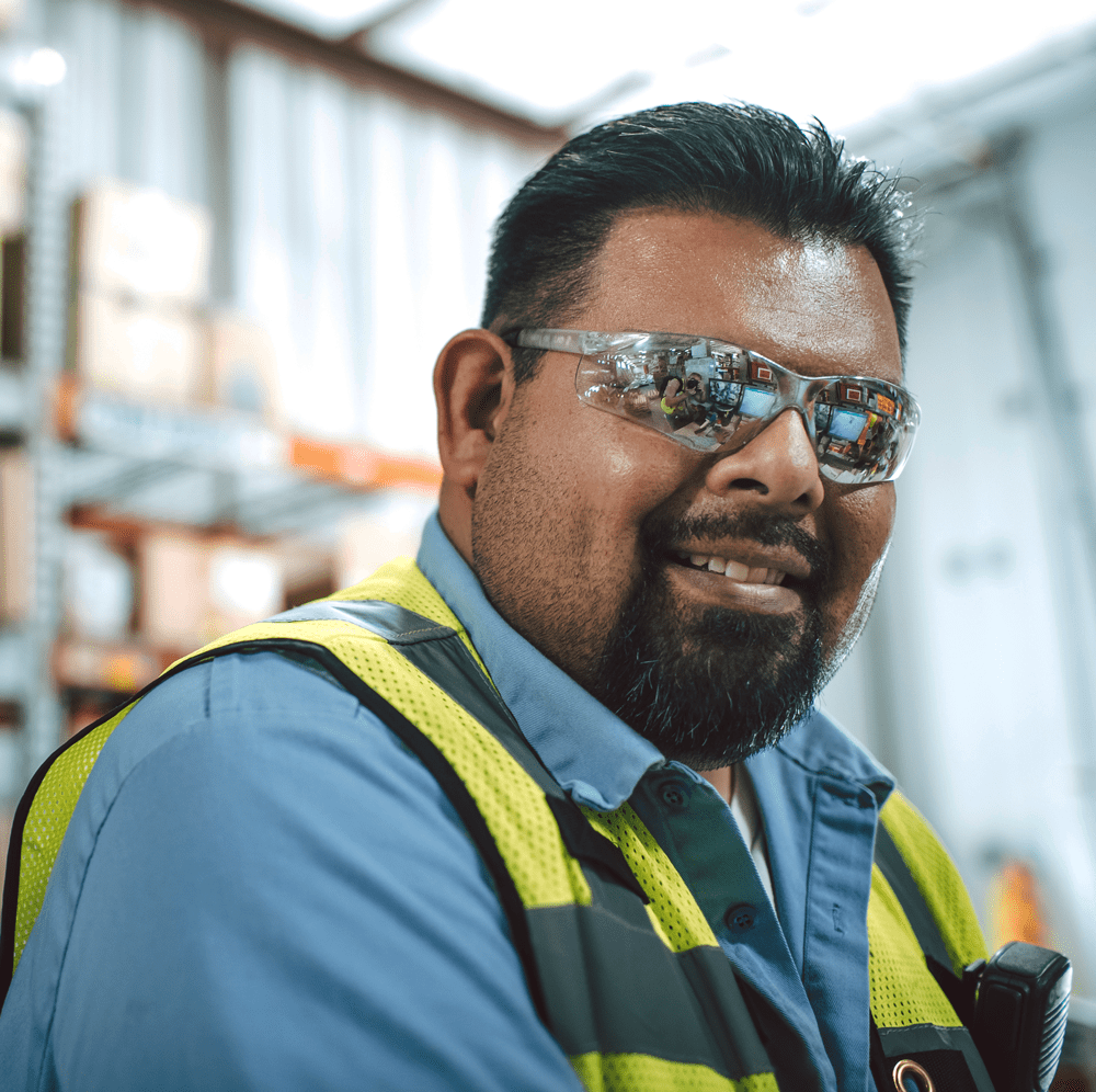 Man with black hair and beard in a production facility wearing a yellow vest and smiling