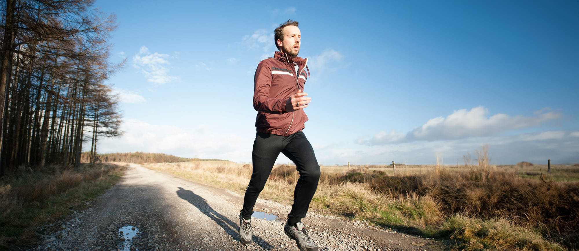 White male in athletic clothing mid stride on a midwest dirt road with a field to his right and a line of pine trees to his left.