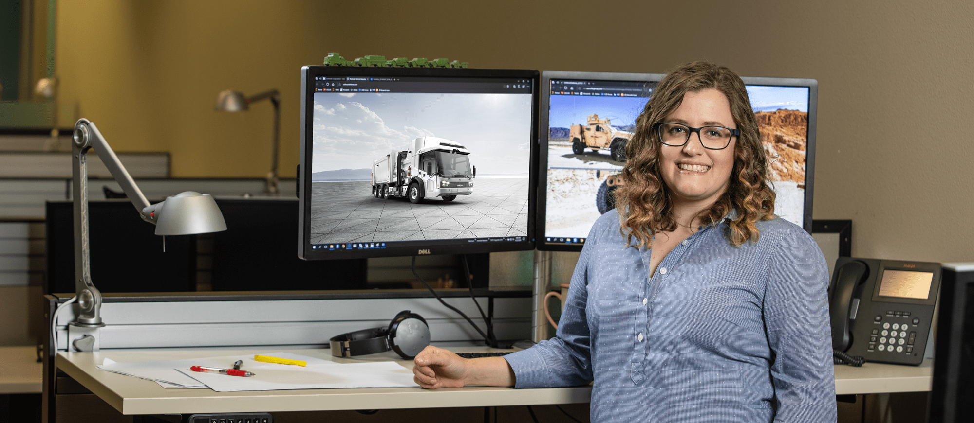 Amanda Miller wearing a blue collared shirt standing in front of a desk and two monitors showing vehicles