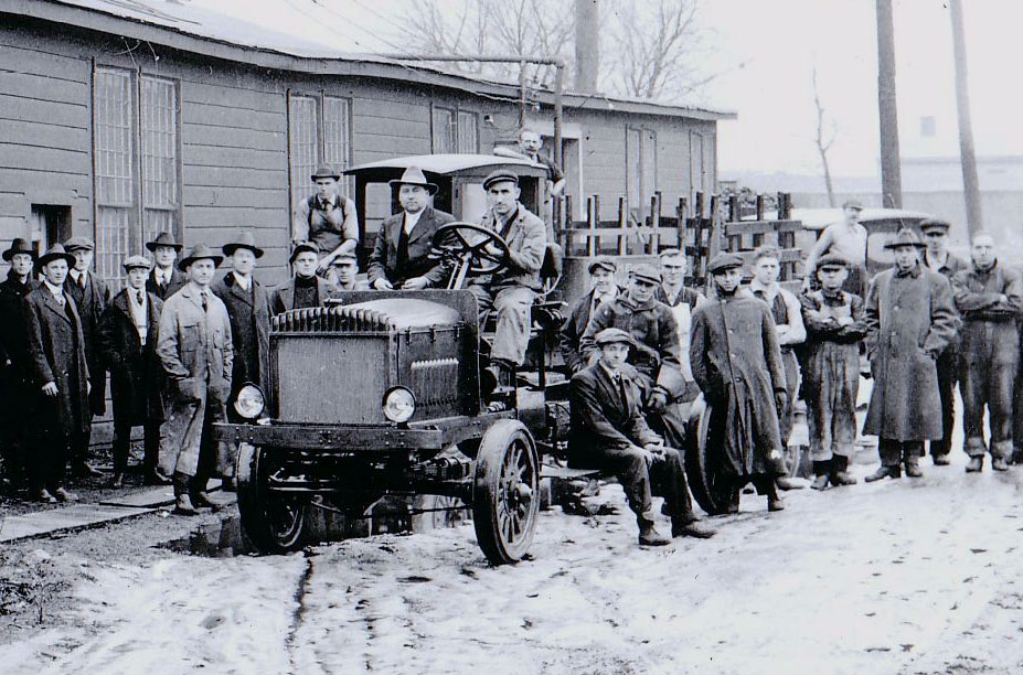 A group of people standing around a truck from the early 1900's