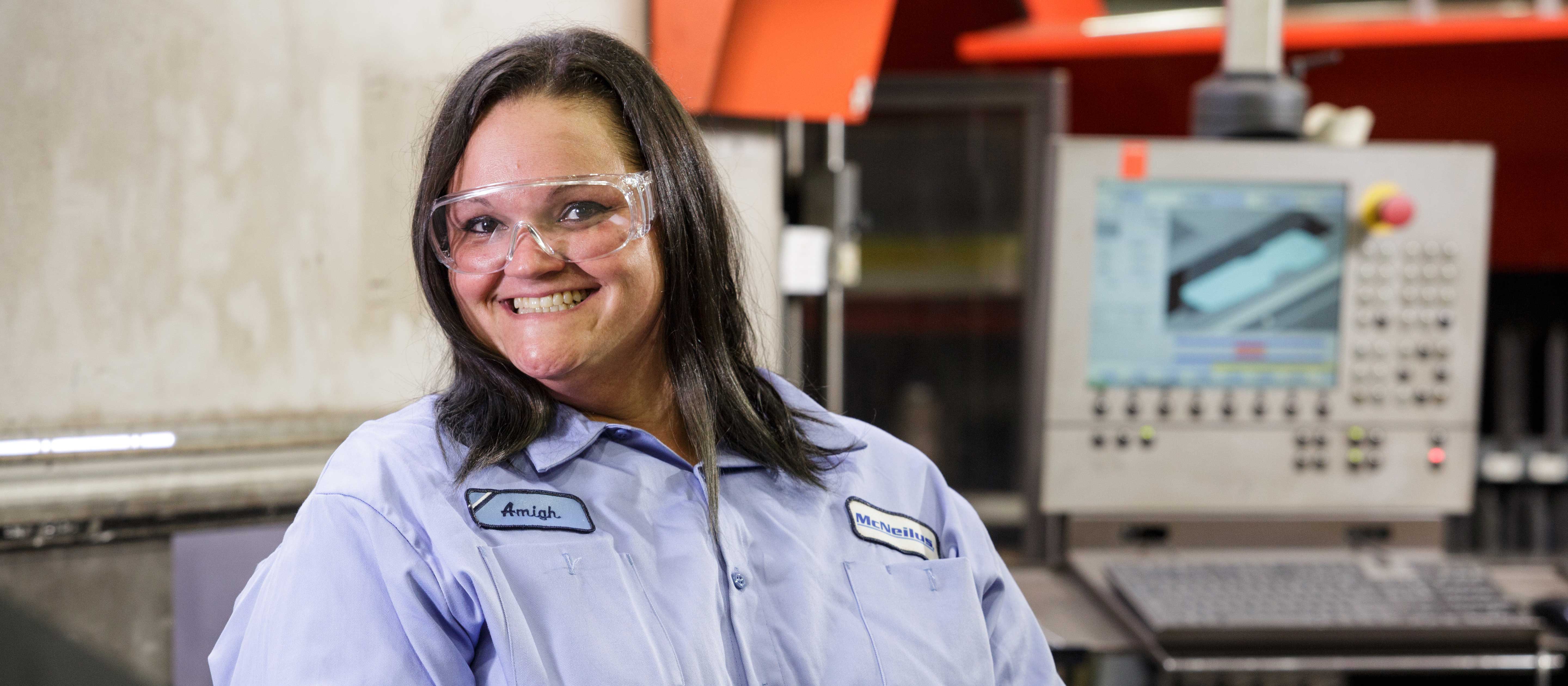 Woman with dark brown hair standing in a production facility smiling in a blue button up McNeilus shirt