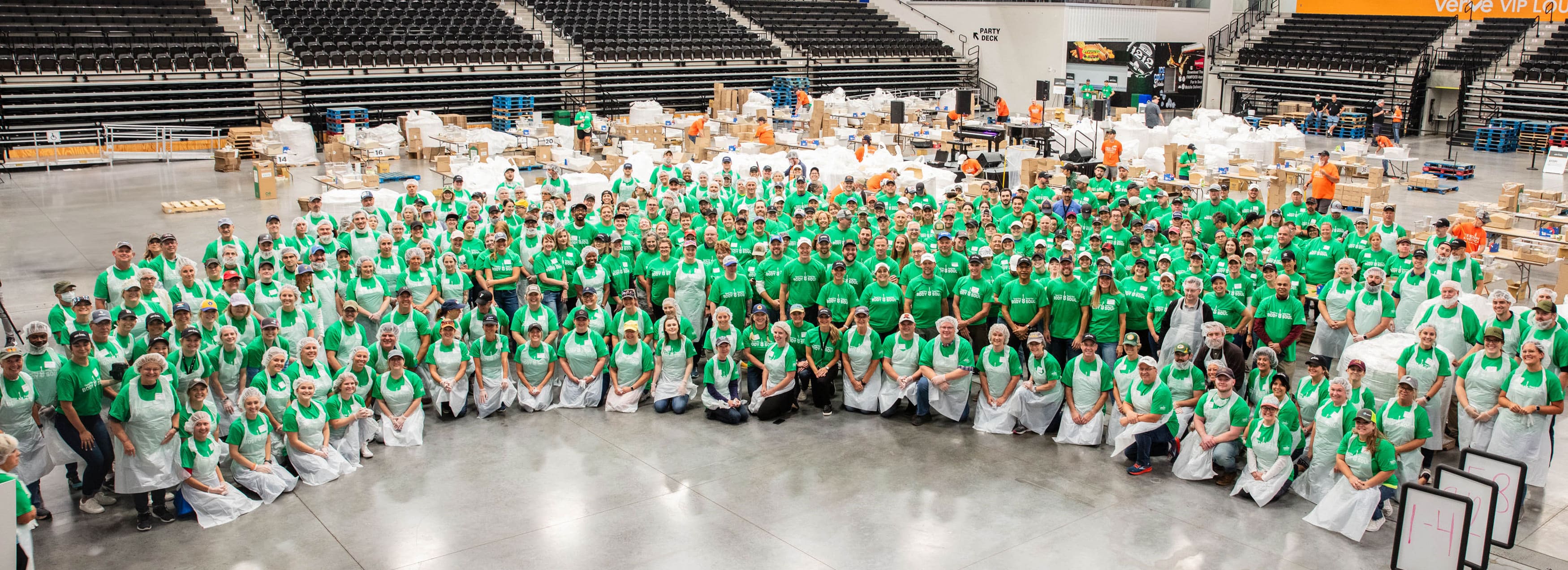 Large group of volunteers wearing green shirts inside a building getting ready to pack rice for Feeding America