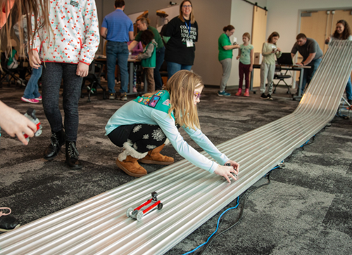Female child girl scout with blonde hair and glasses wearing a green girl scout sash playing with a toy vehicle model on a track