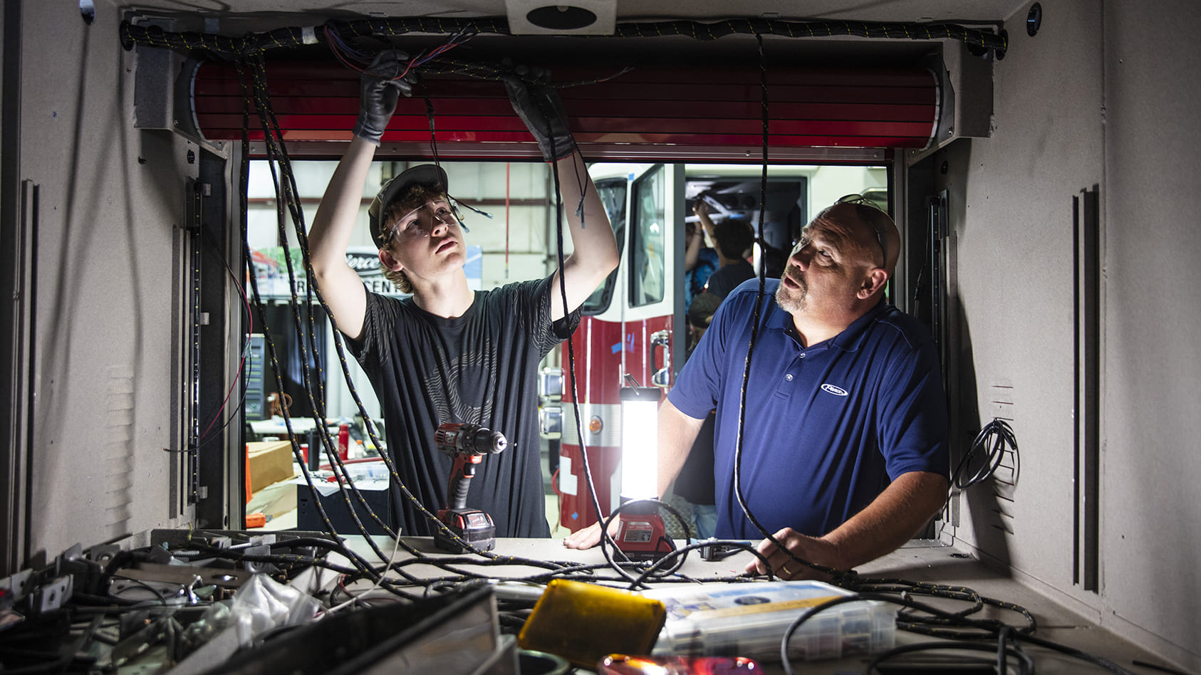 A middle aged male helping a younger male work on electrical on the inside of a truck cab 