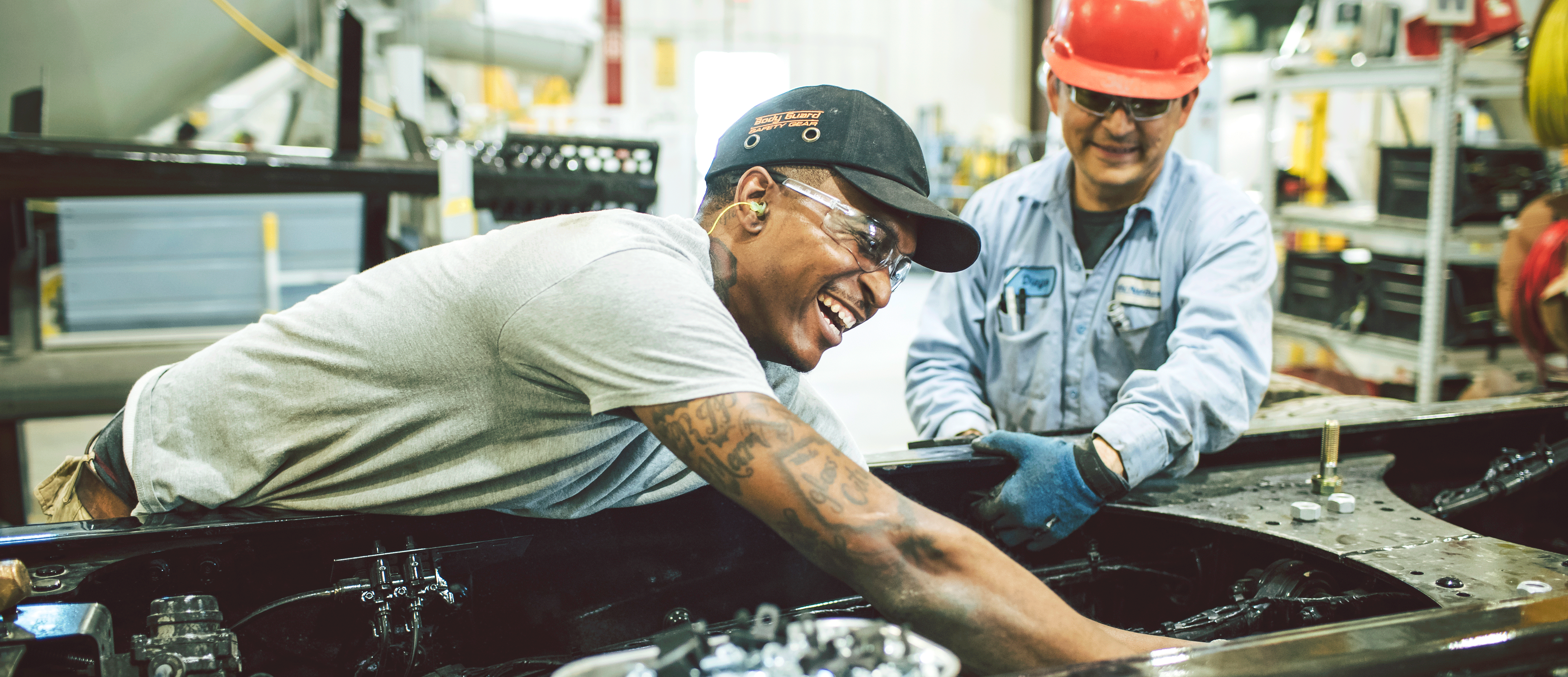 Two men wearing safety gear and smiling while working on an Oshkosh product in a manufacturing facility 